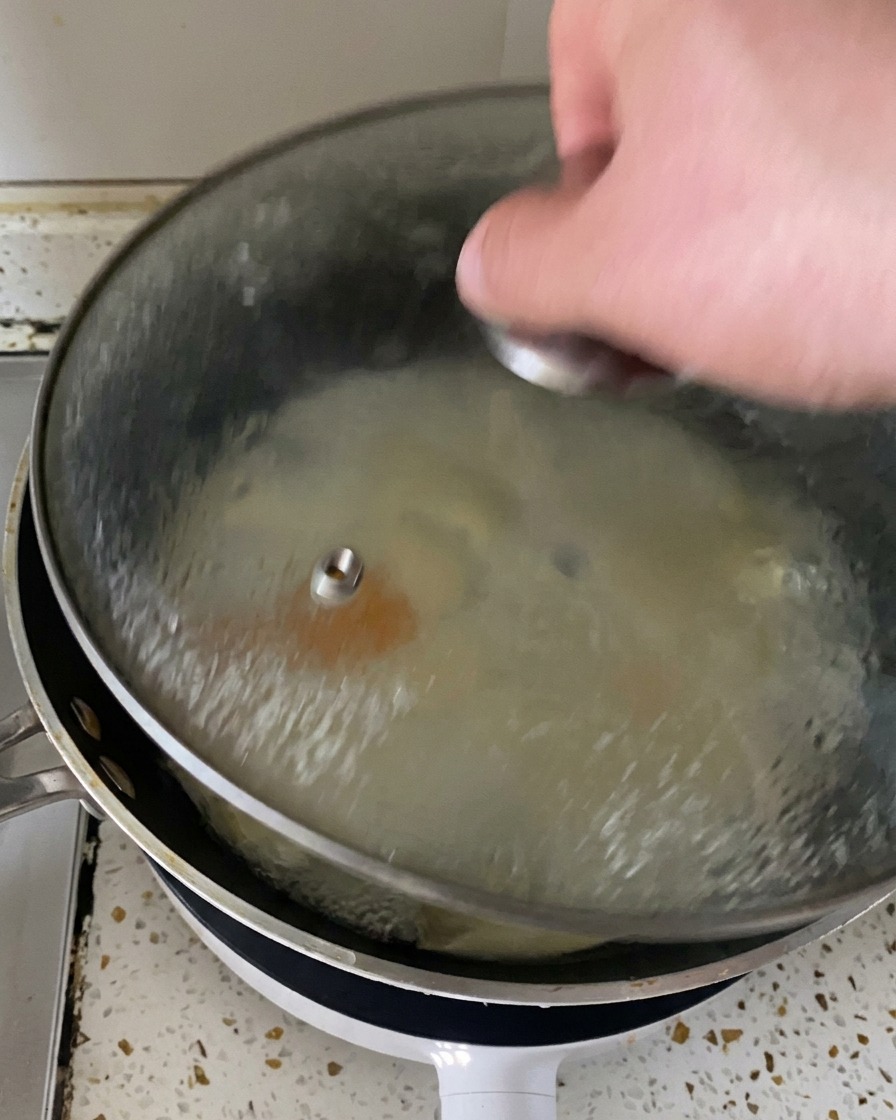 フライパンからガラスの蓋を持ち上げると、湯気とともにチーズが溶けた完璧な目玉焼きが現れる様子。