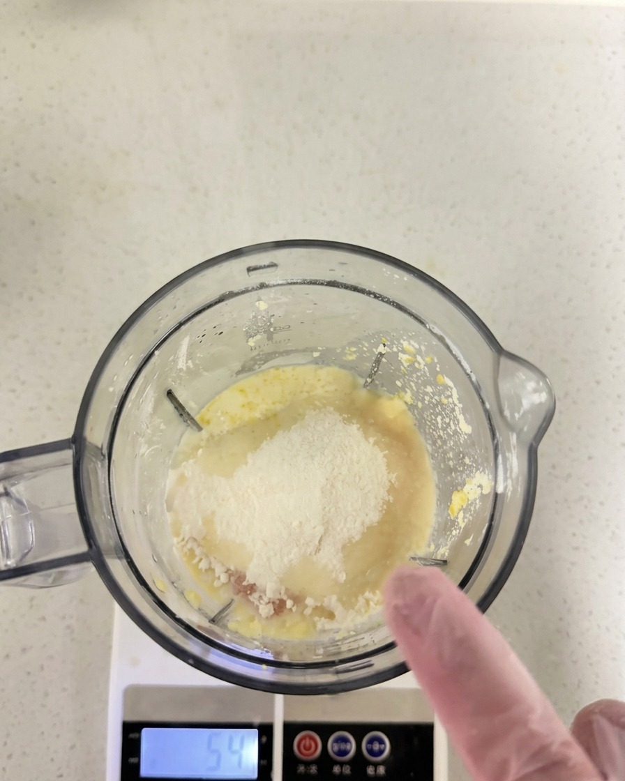 キッチンスケールにのせたミキサーの容器の中で、卵黄などの上に粉類が追加されている様子。