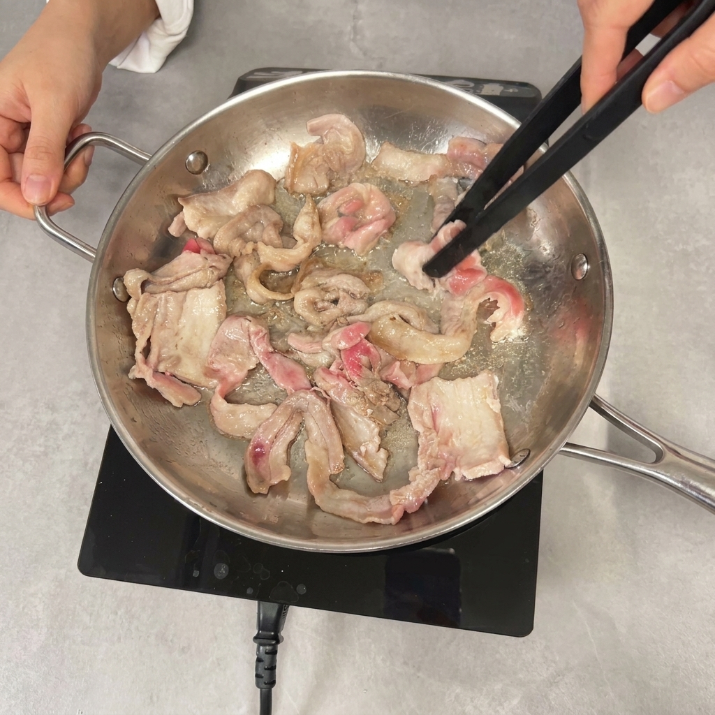 黒いトングを使って、少し焼き色のついた豚バラ肉をフライパンで炒め合わせている様子。
