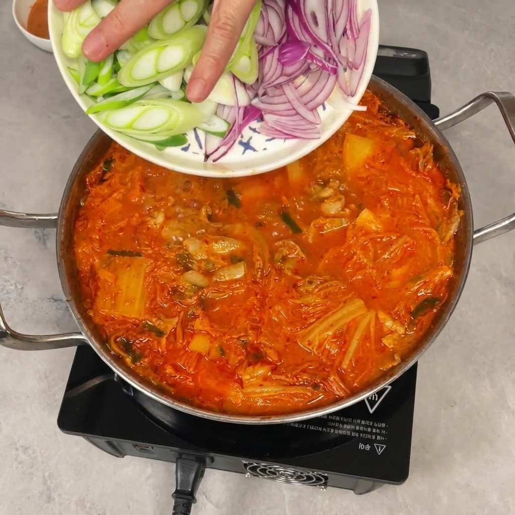 スライスした長ねぎと紫玉ねぎを、お皿から煮立っている赤いキムチチゲの鍋に入れている様子。