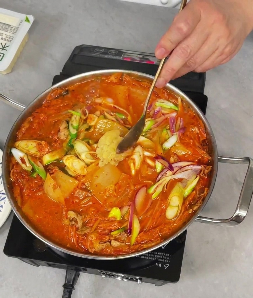 グツグツ煮立つ赤いキムチチゲに、おろしにんにくをスプーン一杯加えている様子。