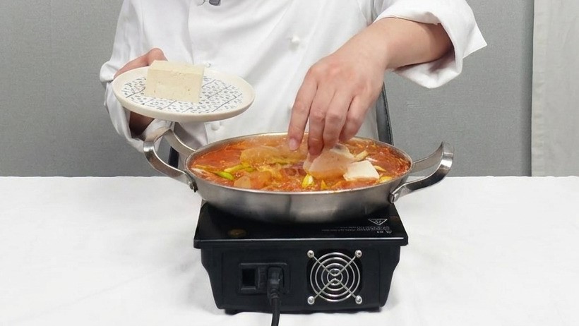 シェフが、四角く切った白い豆腐を煮立っている赤いキムチチゲの鍋にそっと並べている様子。