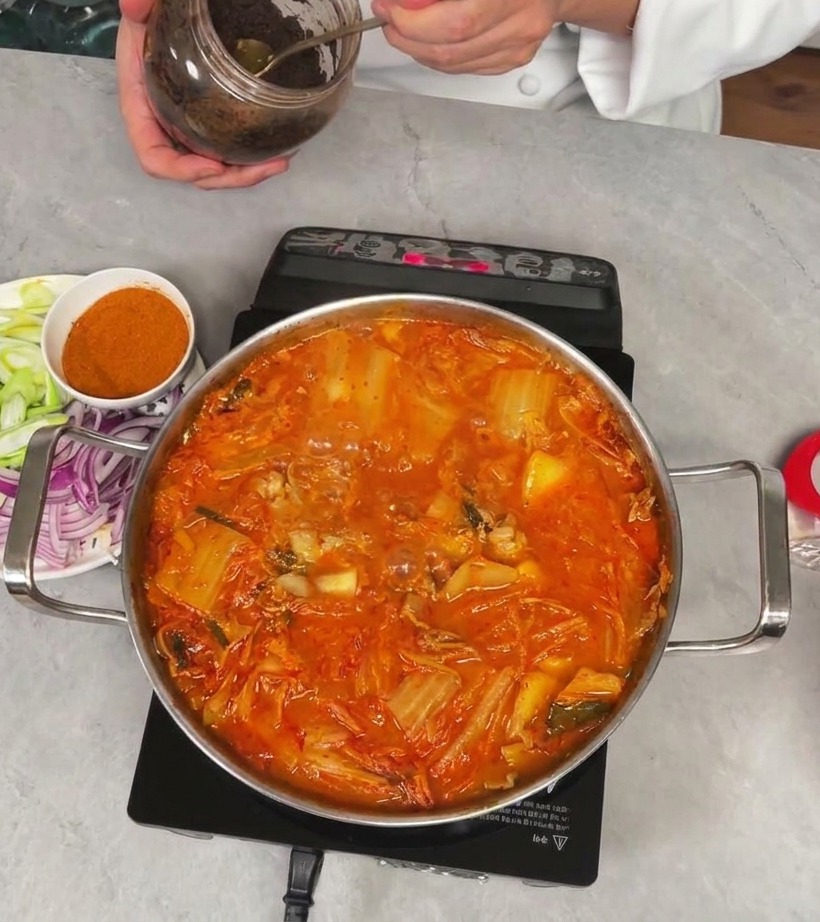 シェフが瓶からテンジャン（韓国味噌）をスプーンですくい、キムチチゲに加えようとしている様子。