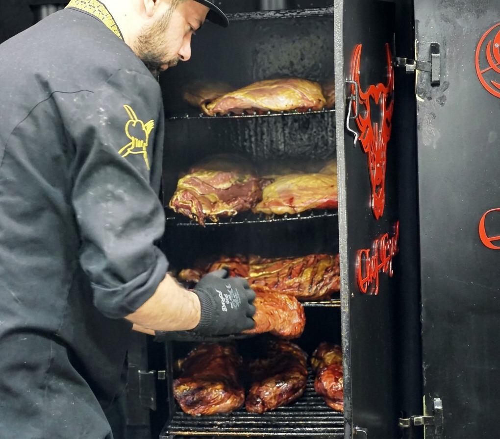 シェフが多段式の肉用スモーカーから、黒い層(バーク)が形成された大きな燻製肉を取り出している様子