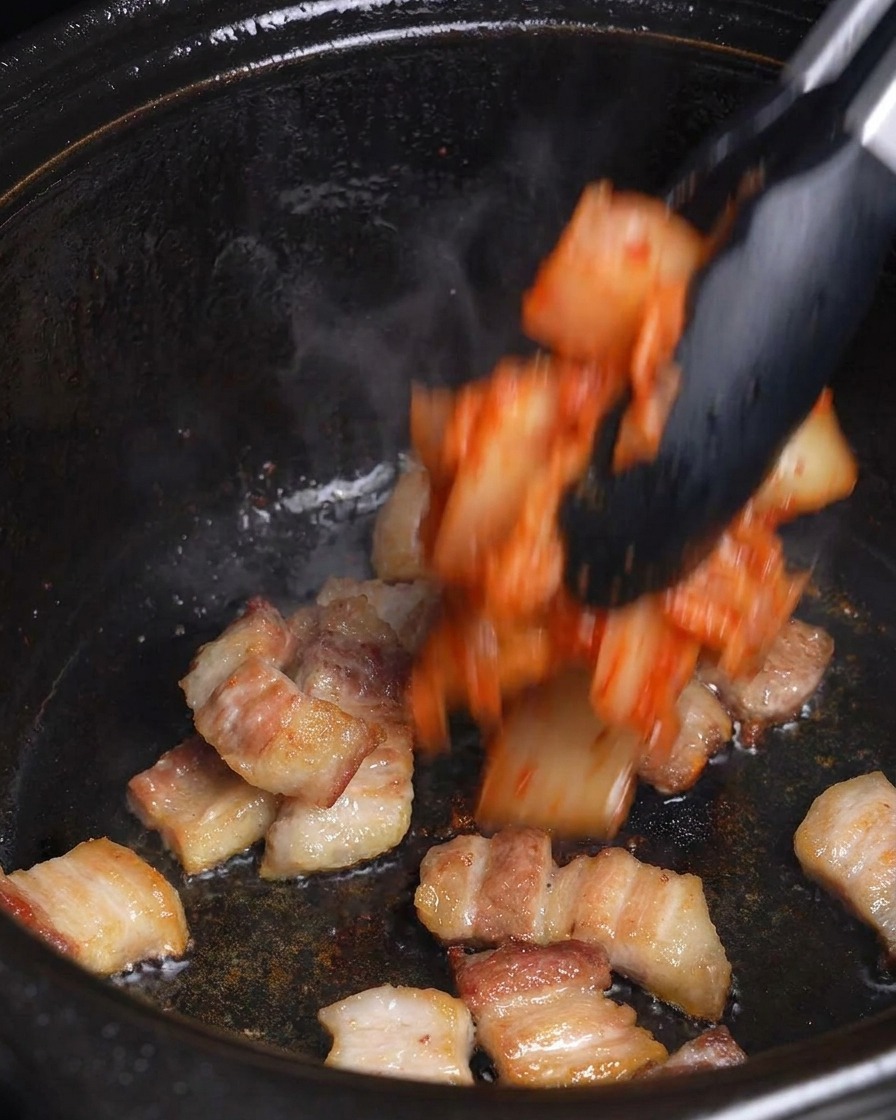 こんがり焼けた豚バラ肉の入った黒い鍋に、刻んだキムチを加えている様子。