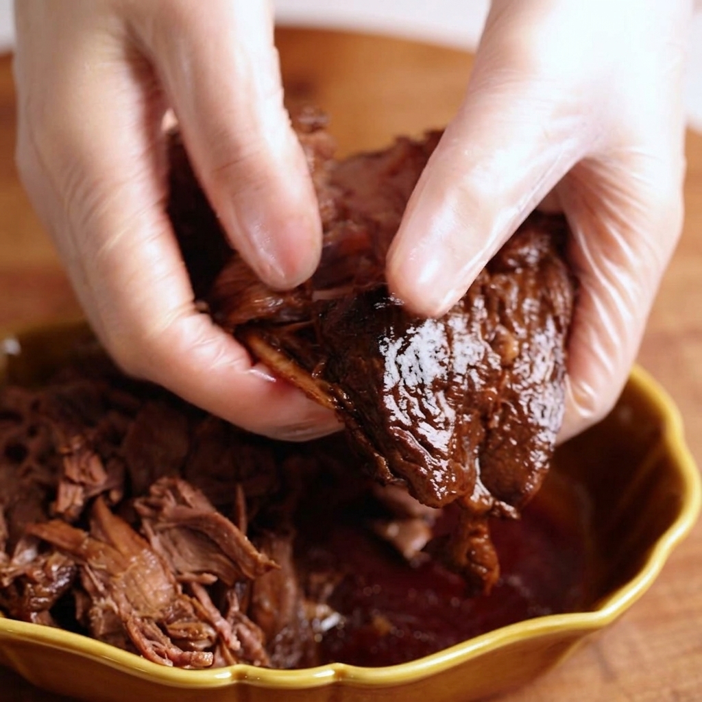 ビニール手袋をした手で、柔らかく煮上がった牛肉を粗くほぐしている様子。