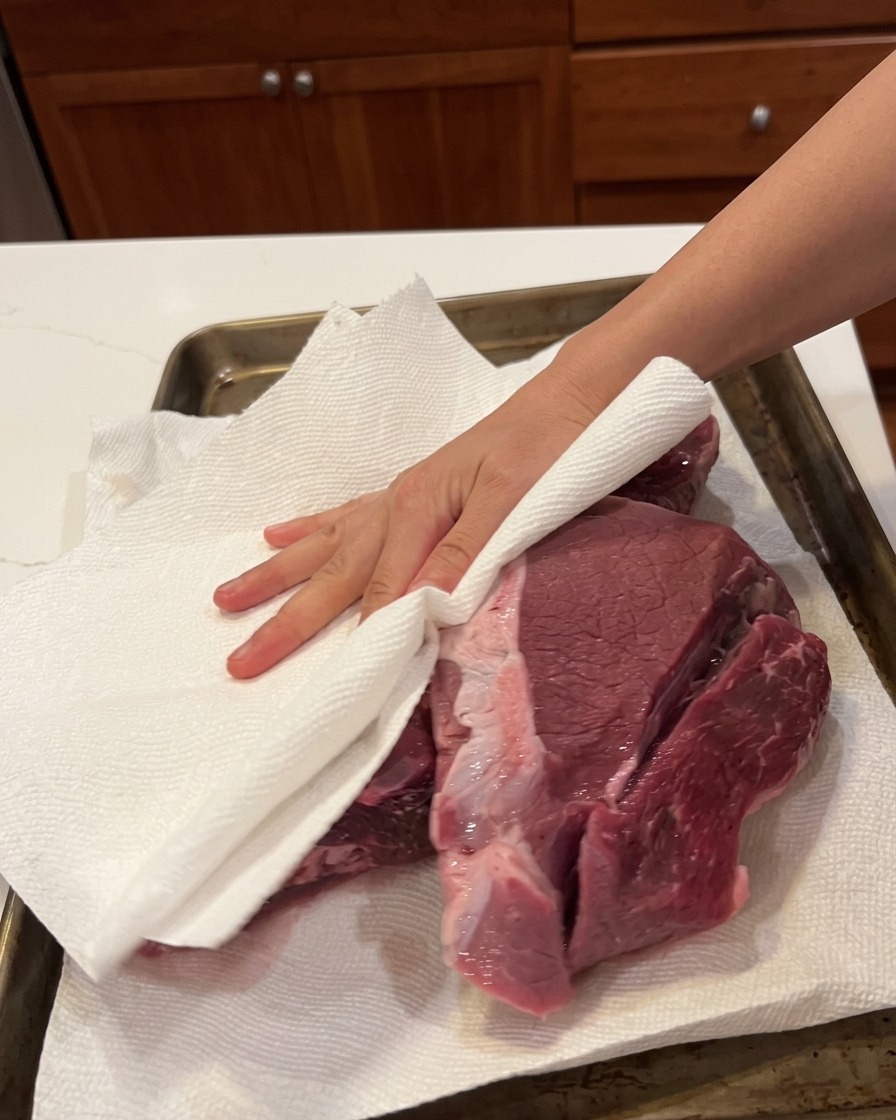 天板の上で、キッチンペーパーを使って大きな塊の牛肉の水気を丁寧に拭き取っている様子。
