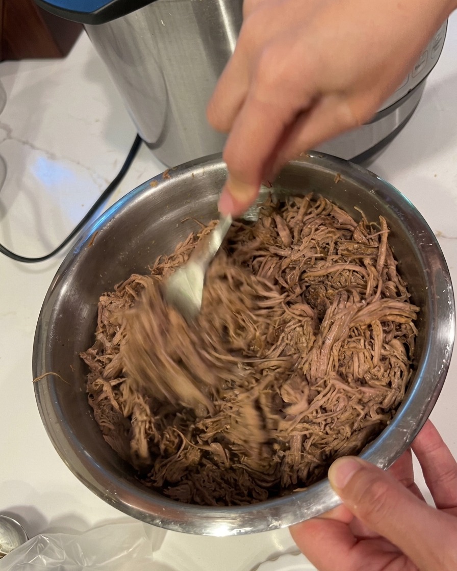 ステンレスボウルの中で、2本のフォークを使って柔らかい肉を細かくほぐしている様子。