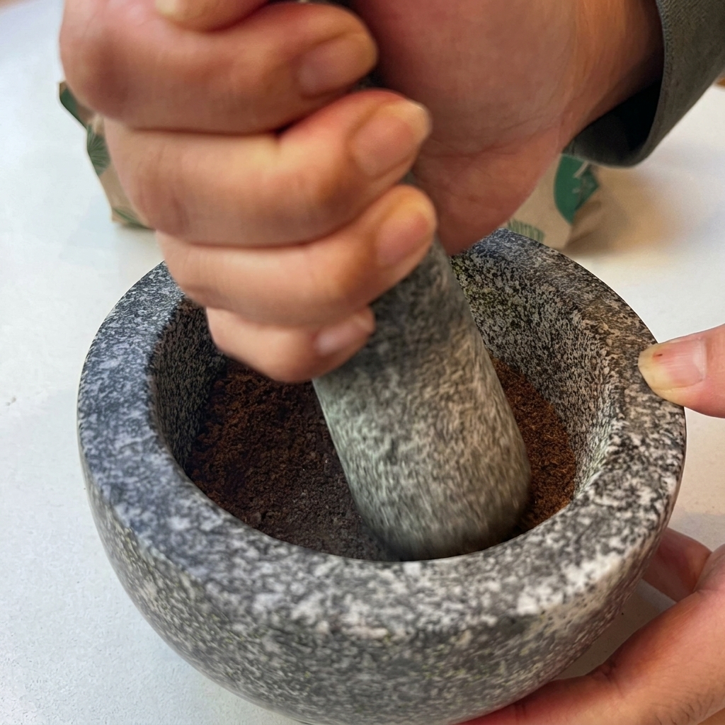 石のすり鉢の中で、煎りたてのスパイスをすりこぎで細かい茶色の粉末に挽いている手元の様子。