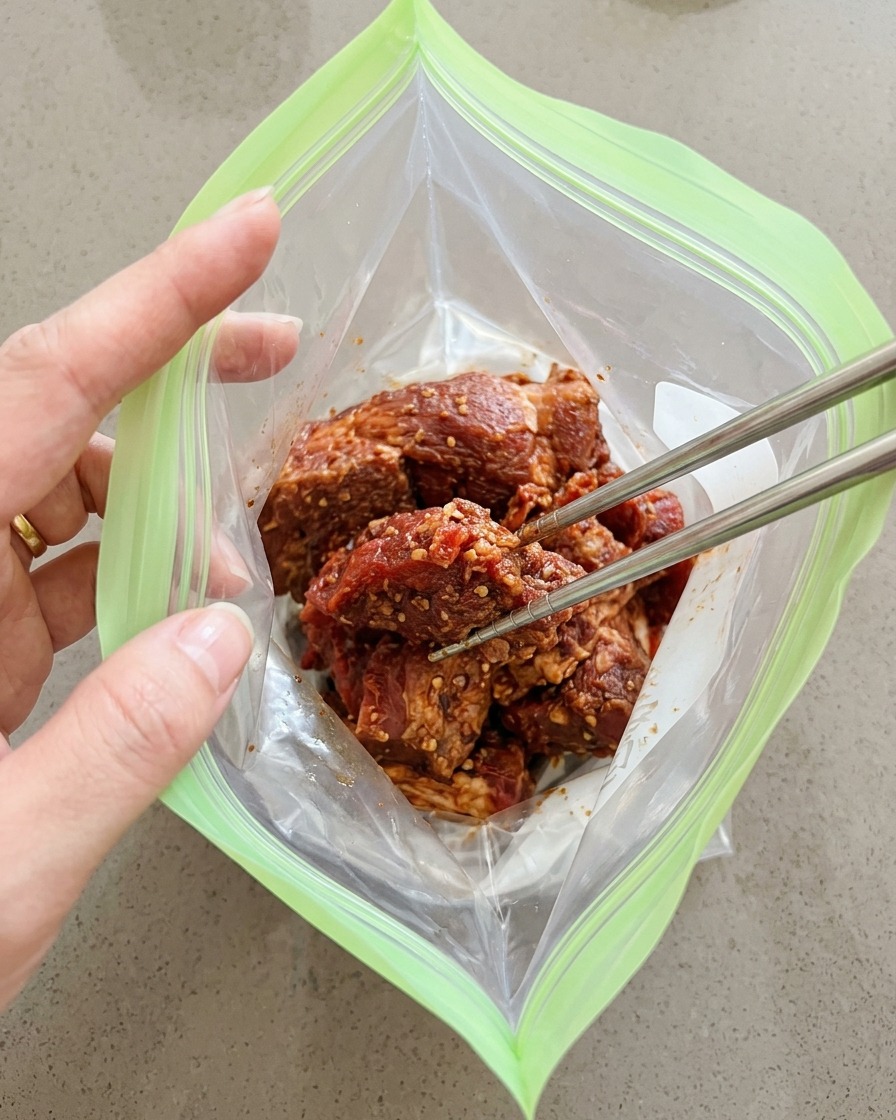 味付けした豚肉を、金属製の箸を使って緑色のジップ付き保存袋に入れている様子。