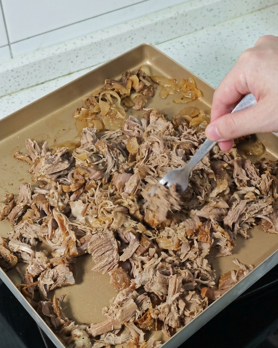 バットの上で、熱々の煮込み肉をフォークを使って細かくほぐしている様子。