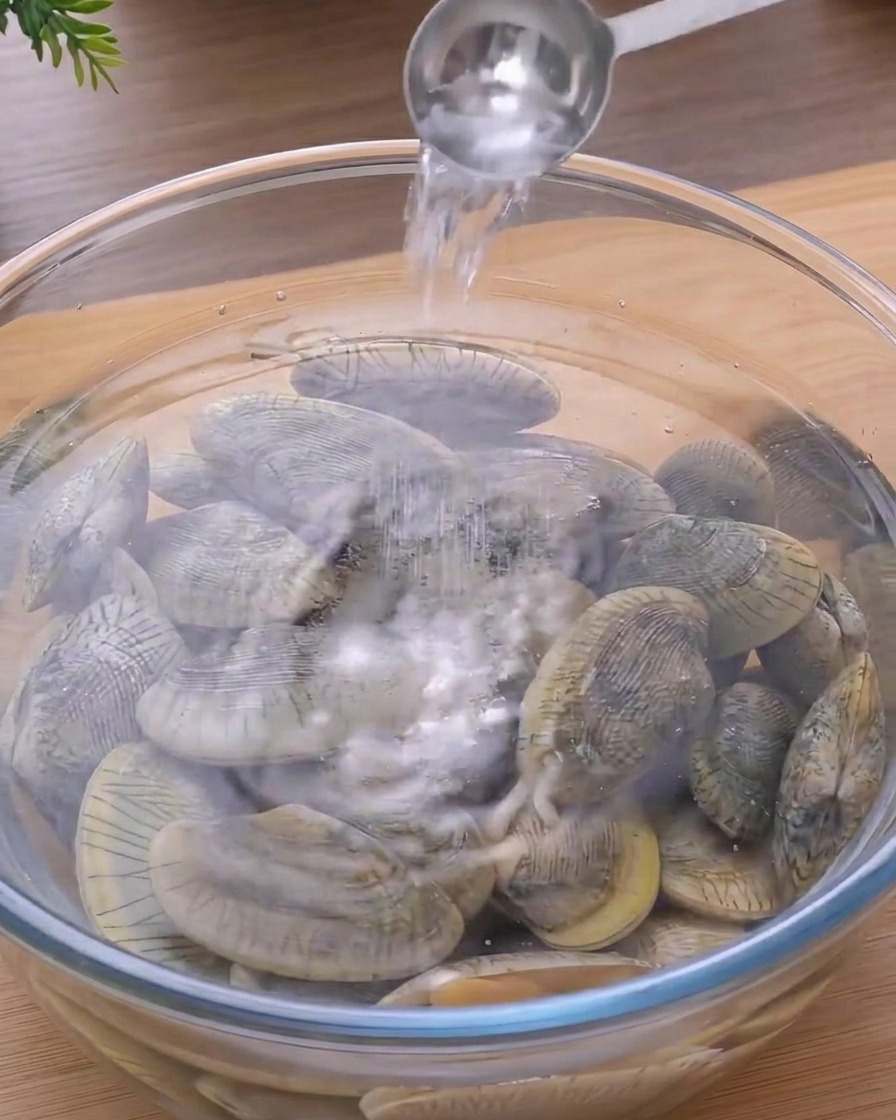 水と新鮮なあさりが入った透明なガラスボウルに、スプーンで塩を入れている様子。