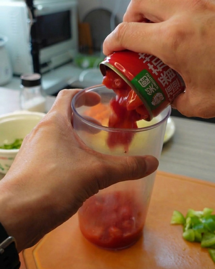 カットされたトマト缶の中身を、縦長の透明なプラスチックのブレンダー容器に注いでいる様子。