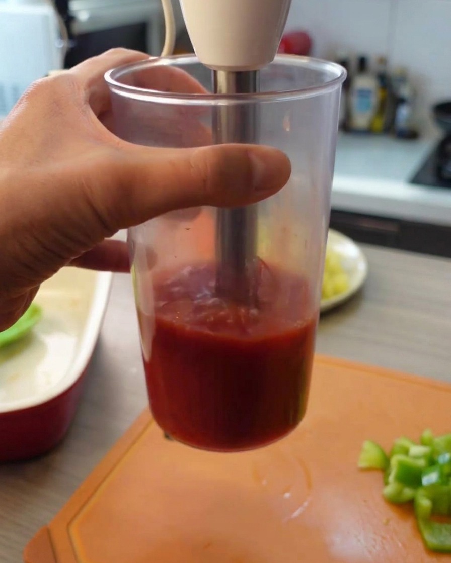 縦長の透明なプラスチック容器の中で、ハンドブレンダーを使ってトマト缶をピュレ状にしている様子。