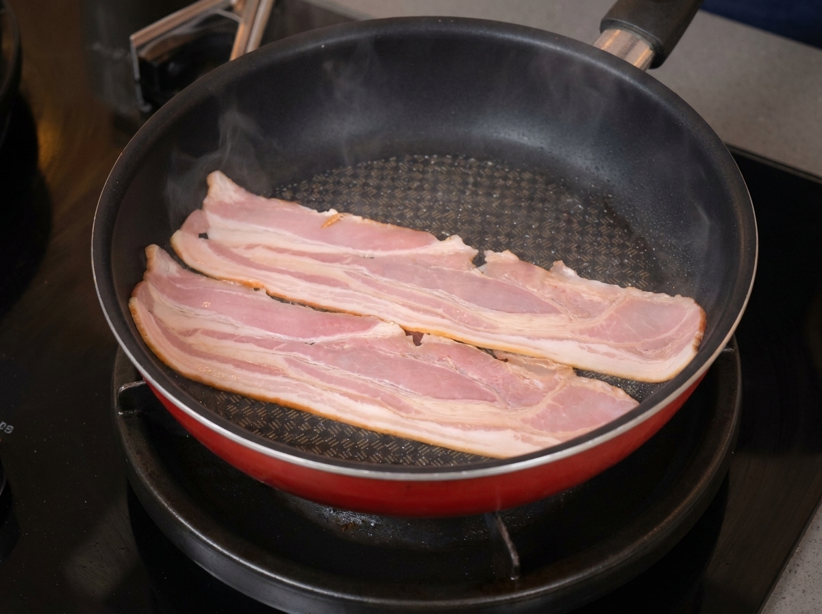フライパンの中で、2枚の長いベーコンが脂を出しながら焼き色がついている様子。