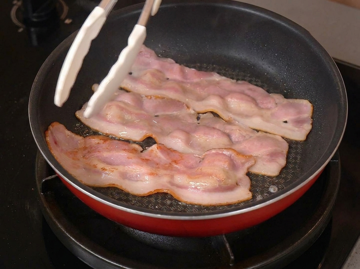 トングを使って、フライパンで香ばしく焼けたベーコンを裏返したり整えたりしている様子。