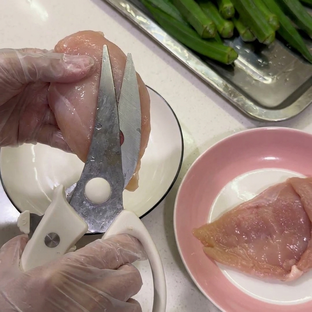 手袋をはめた手で、キッチンバサミを使って生の鶏胸肉から白い脂身を切り取っている様子。