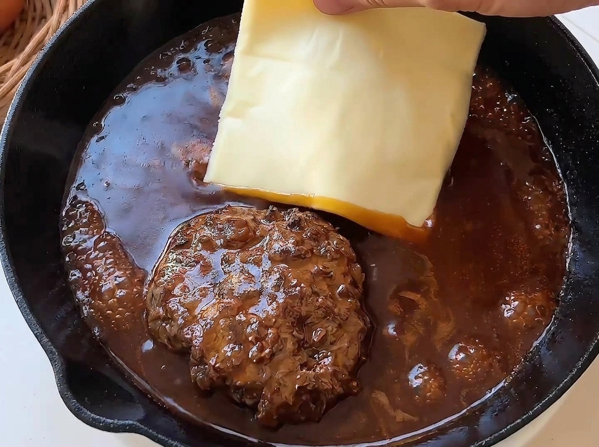 ソースで煮込まれているハンバーグの上に、手でスライスチーズをのせる様子。