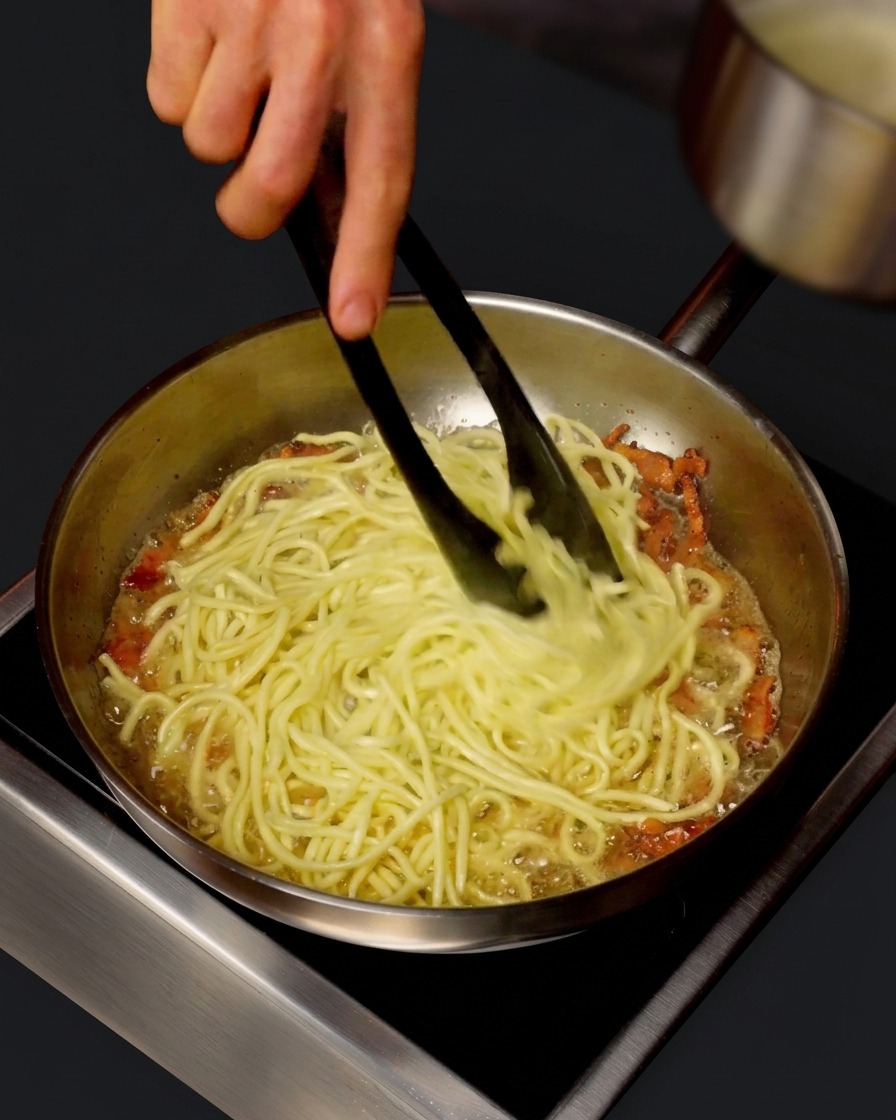 小さな鍋から、スパゲッティとグアンチャーレが入ったフライパンに熱いゆで汁を注ぎ、トングで混ぜ合わせている様子。