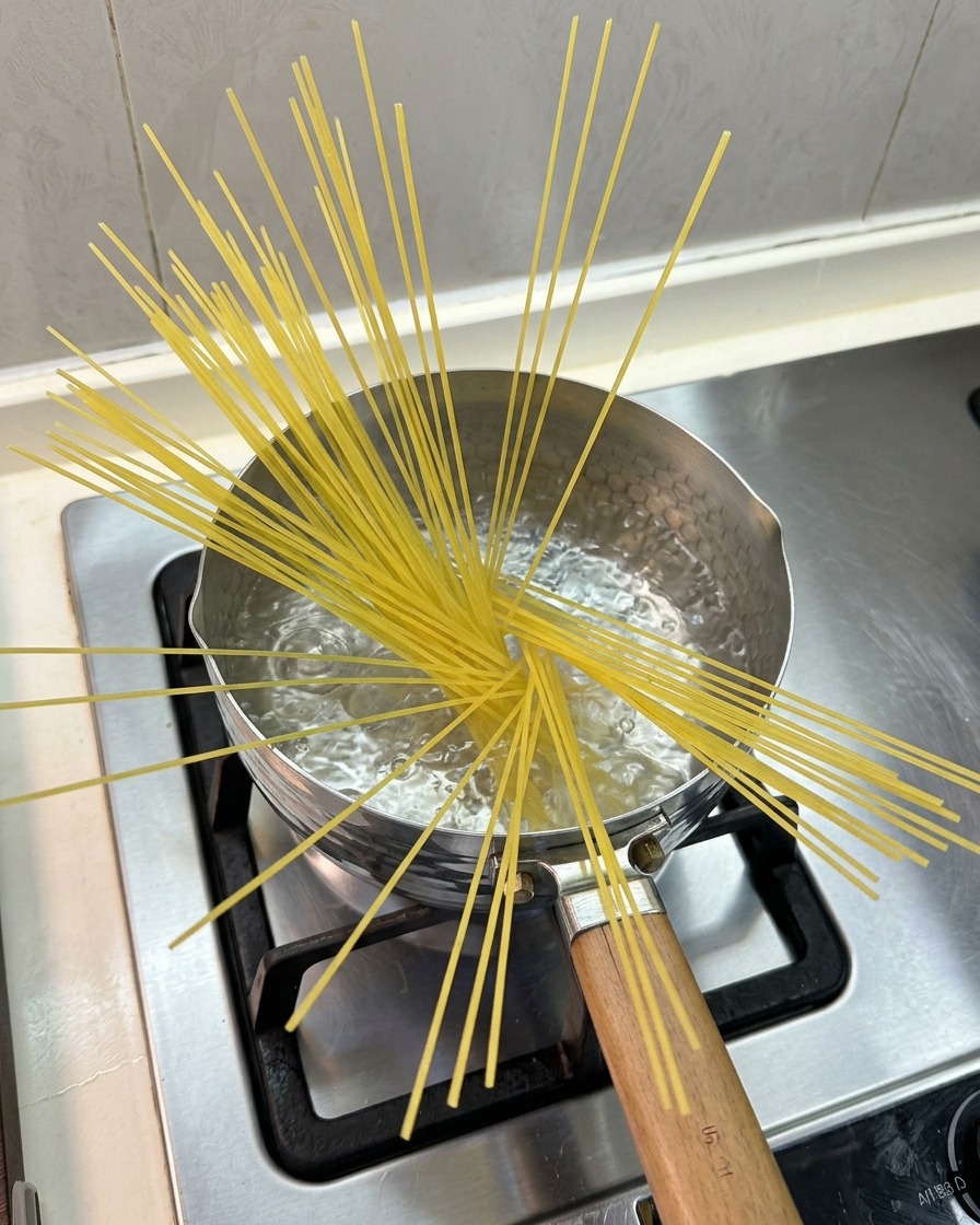ガスコンロの上の小さな鍋で沸騰したお湯に、乾燥したスパゲッティが扇状に広がりながら半分ほど浸かっている様子。