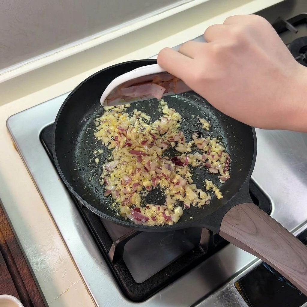 炒めた玉ねぎとにんにくが入ったフライパンに、小さな小皿からスライスしたベーコンを滑り込ませている様子。