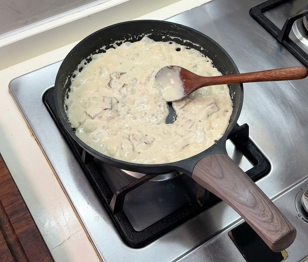 黒いフライパンでとろみのあるクリームソースがグツグツと煮立っており、木製のスプーンでかき混ぜられている様子。
