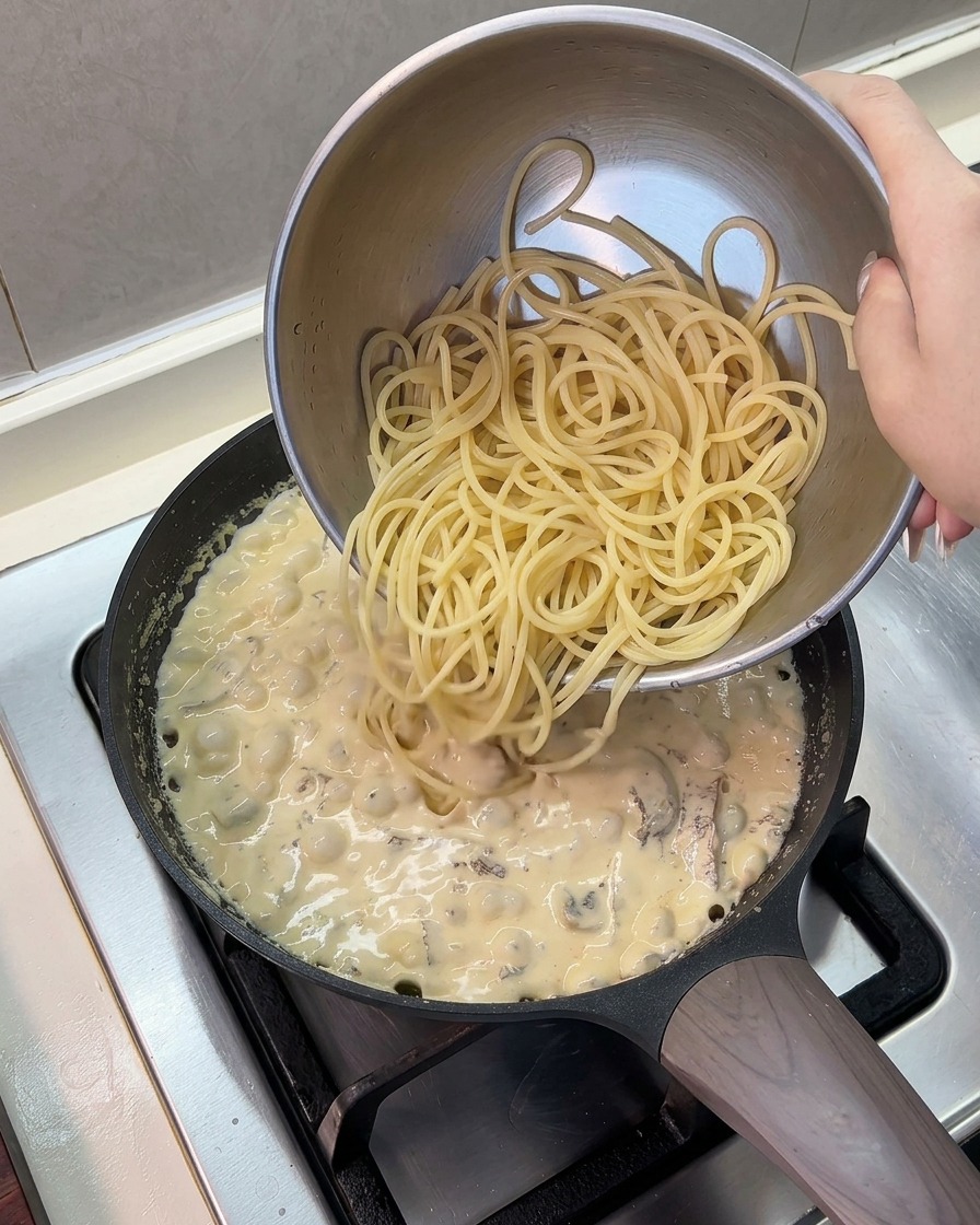 ステンレス製のボウルから茹でたスパゲッティを、煮立っているクリームソースのフライパンに流し入れているところ。