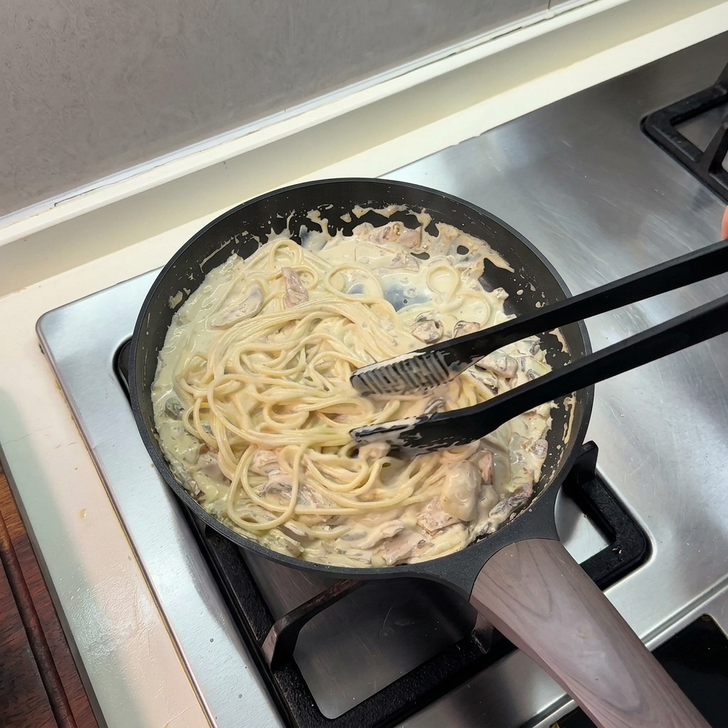 フライパンの中で、黒いキッチントングを使って茹でたスパゲッティと濃厚なクリームソースを絡めている様子。