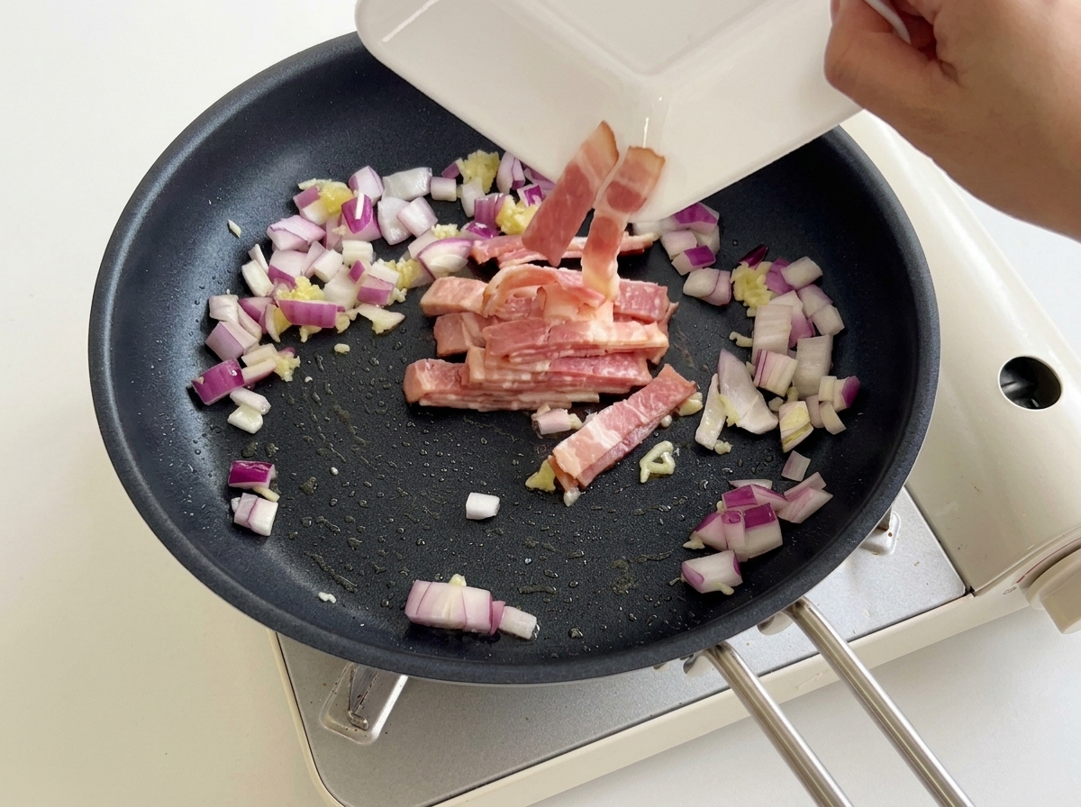 にんにくと玉ねぎが入ったフライパンに、ベーコンを加えて炒める様子。