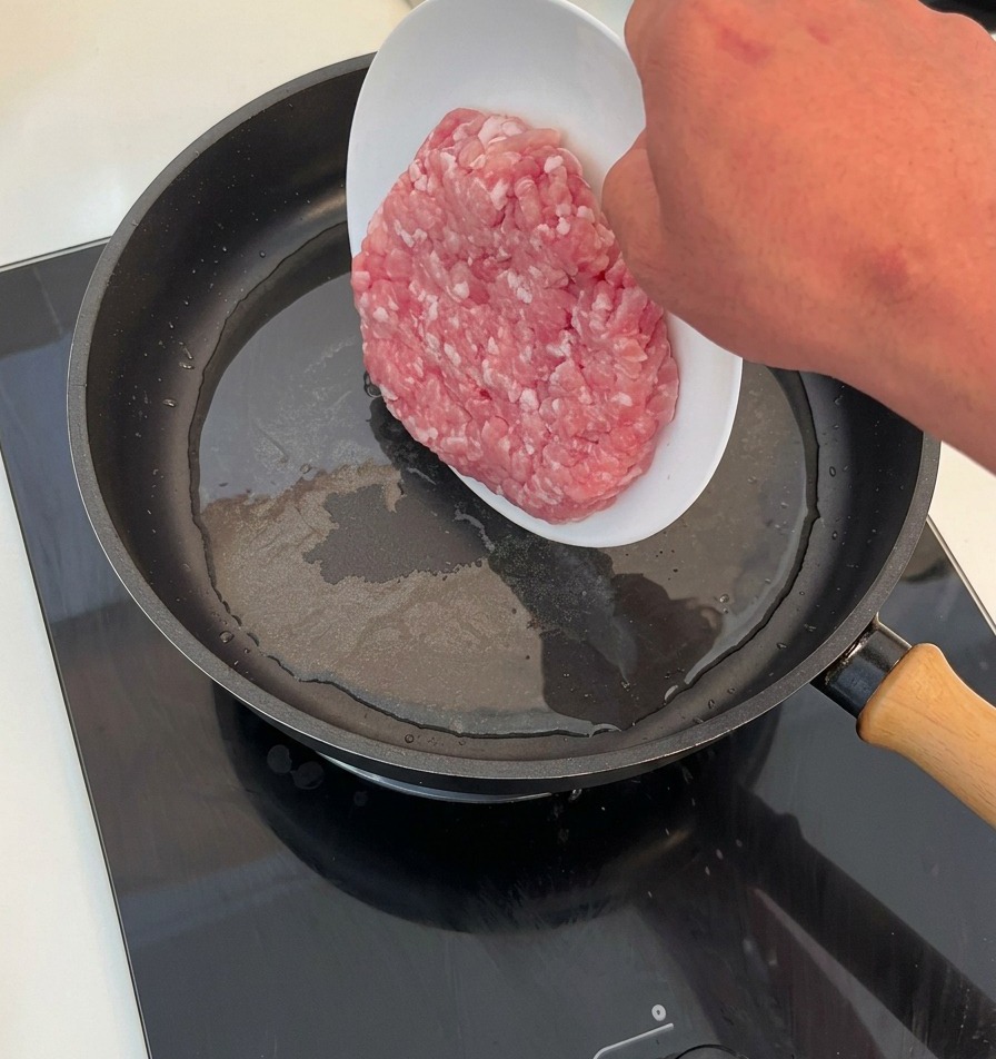 熱したフライパンに、白い皿から生の挽肉をスライドさせて入れている様子。