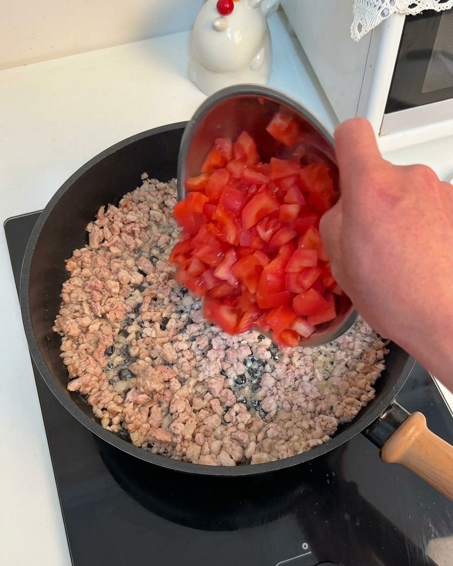 挽肉が焼けているフライパンに、ボウルに入った鮮やかな赤色の角切りトマトを加えている様子。