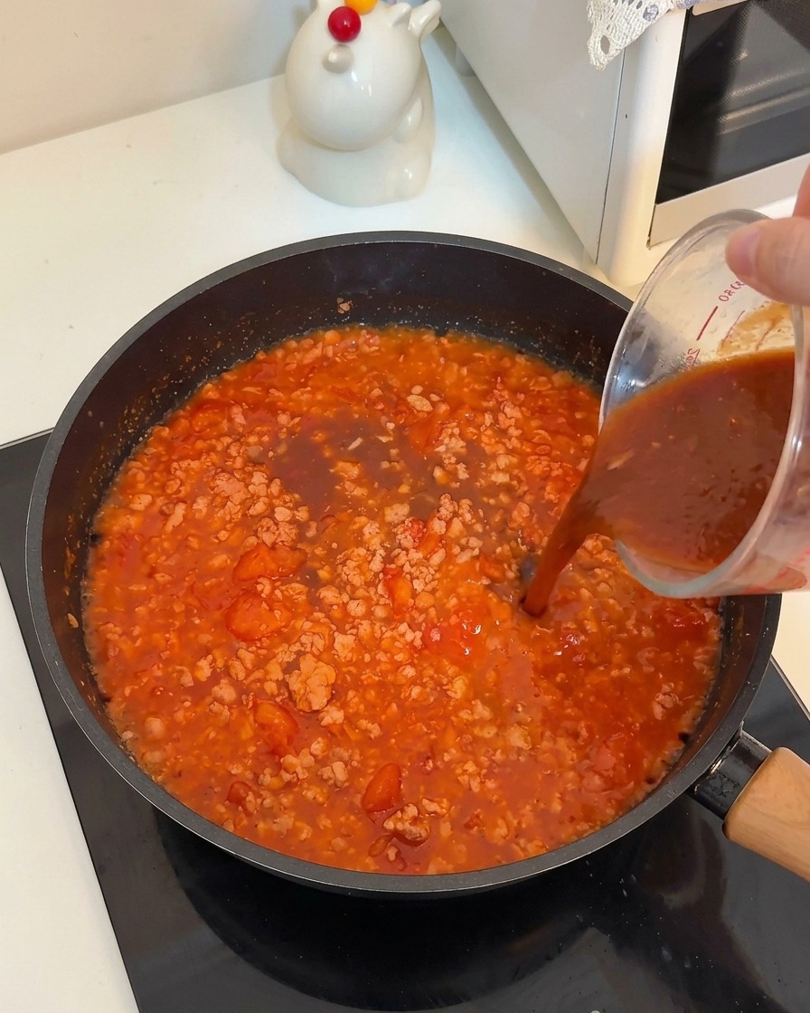 煮込んでいる肉とトマトの中に、計量カップから褐色のソースを注いでいる様子。