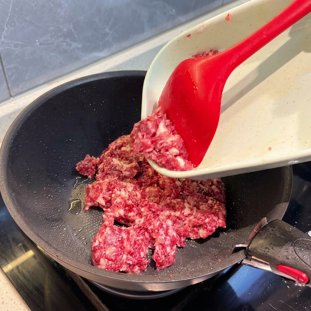 黒いフライパンに、粗挽きにした混合肉を入れ、赤いヘラで広げながら炒めている様子。