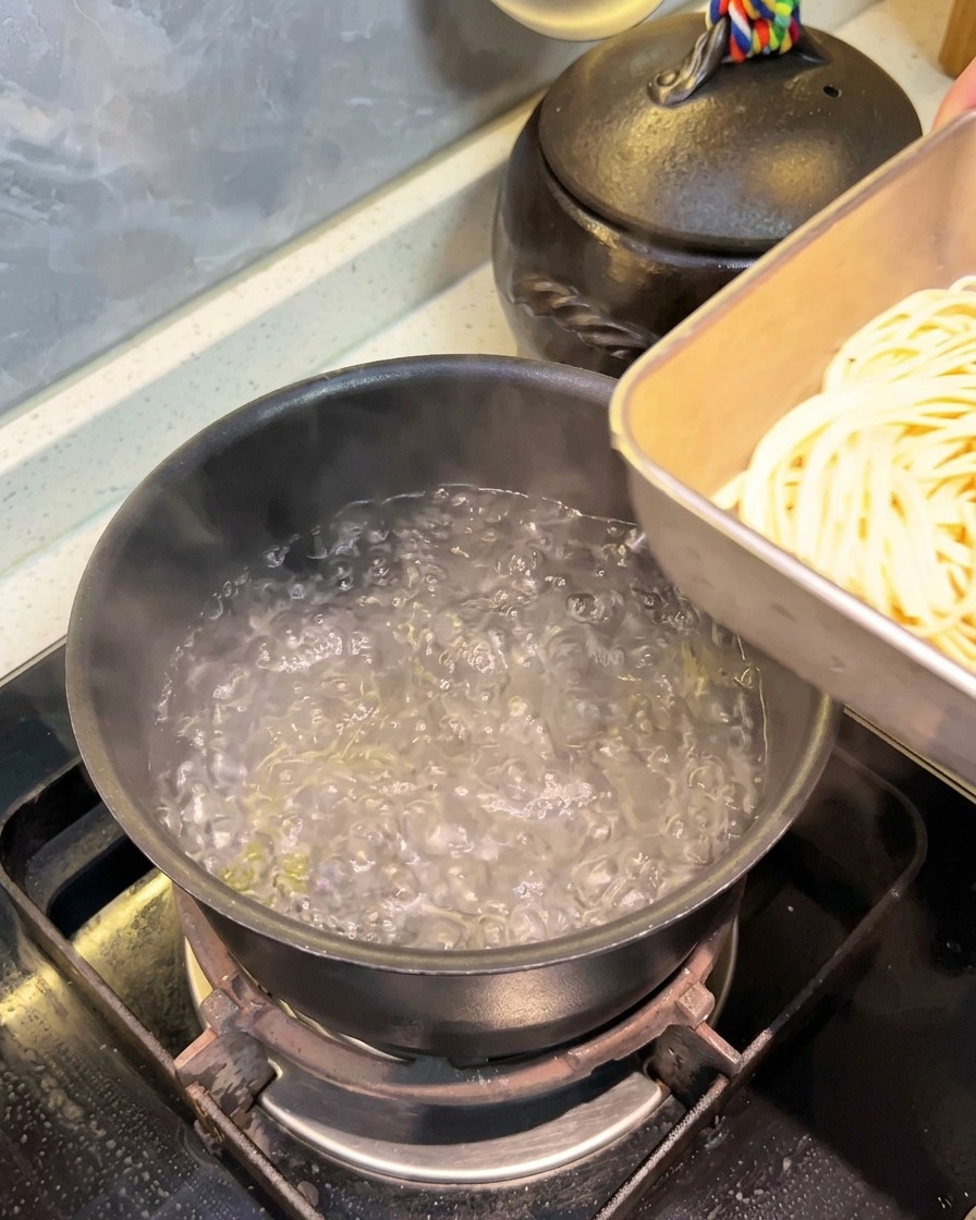 沸騰したお湯が入った黒い鍋に、水に浸しておいたスパゲッティをバットから移し入れている様子。