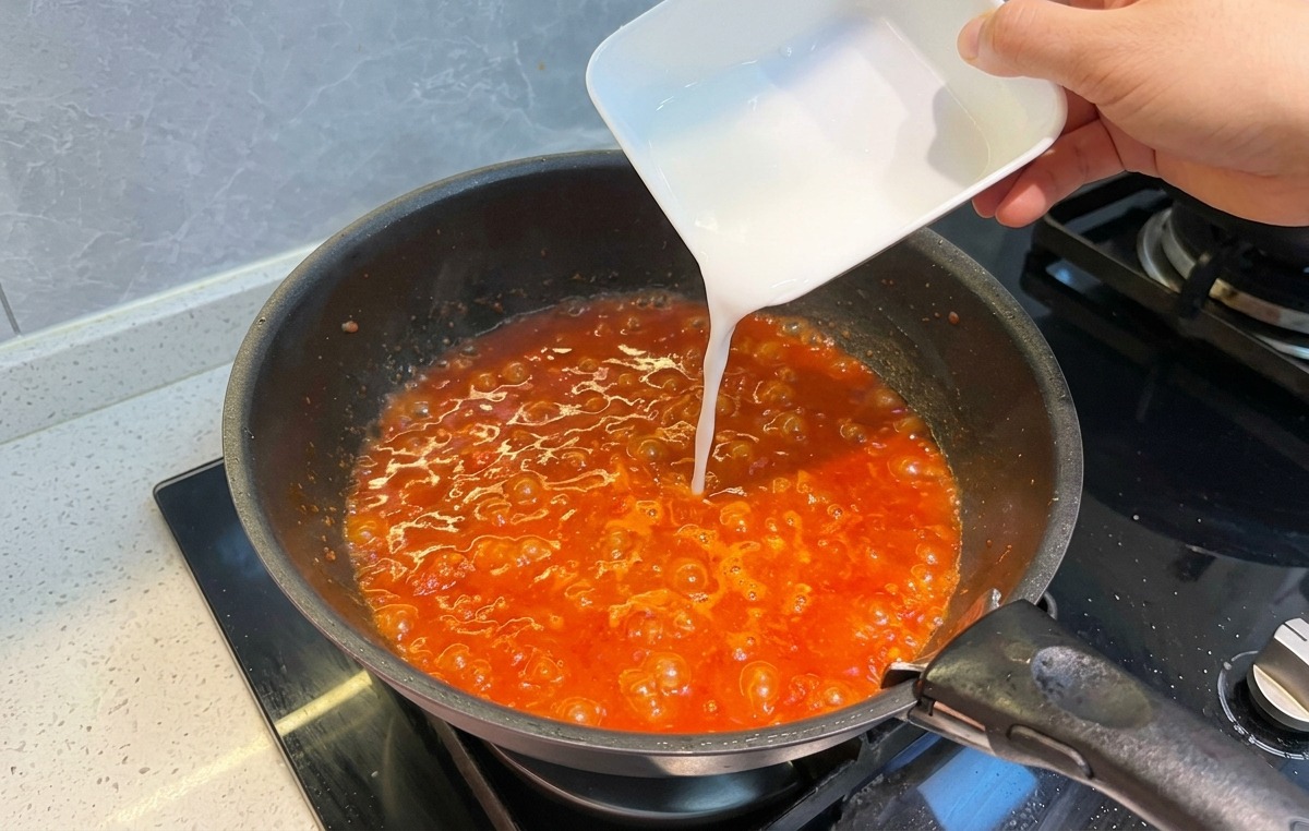 煮え立つ赤いミートソースに、小さな器から白い水溶き片栗粉を回し入れている様子。
