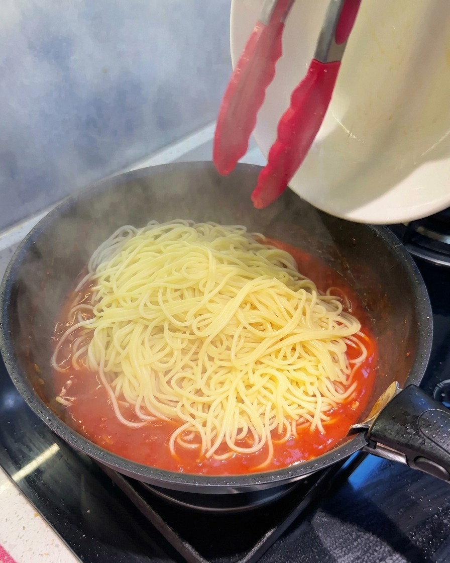 トングを使い、茹で上がったスパゲッティを、煮込まれた厚みのあるミートソースのフライパンへ移している様子。