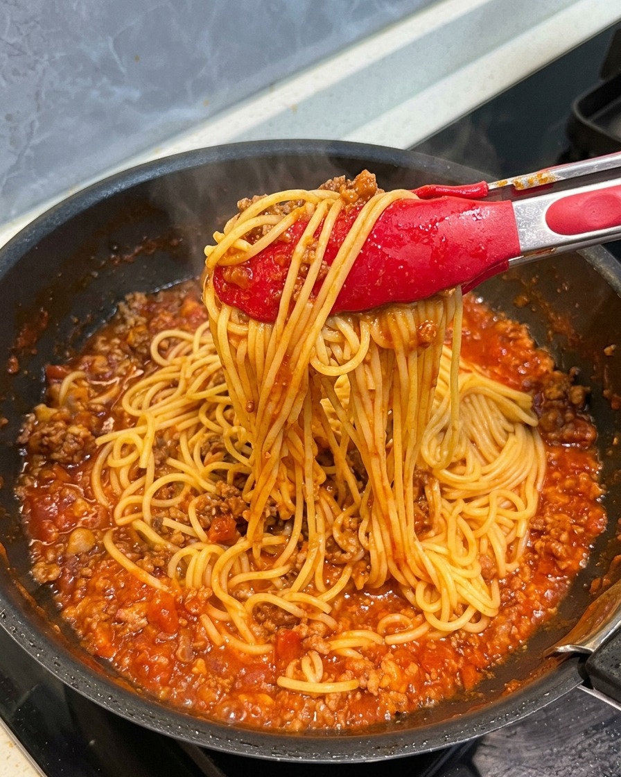 トングでパスタを持ち上げ、濃厚なミートソースが麺の細部までしっかりコーティングされている様子。