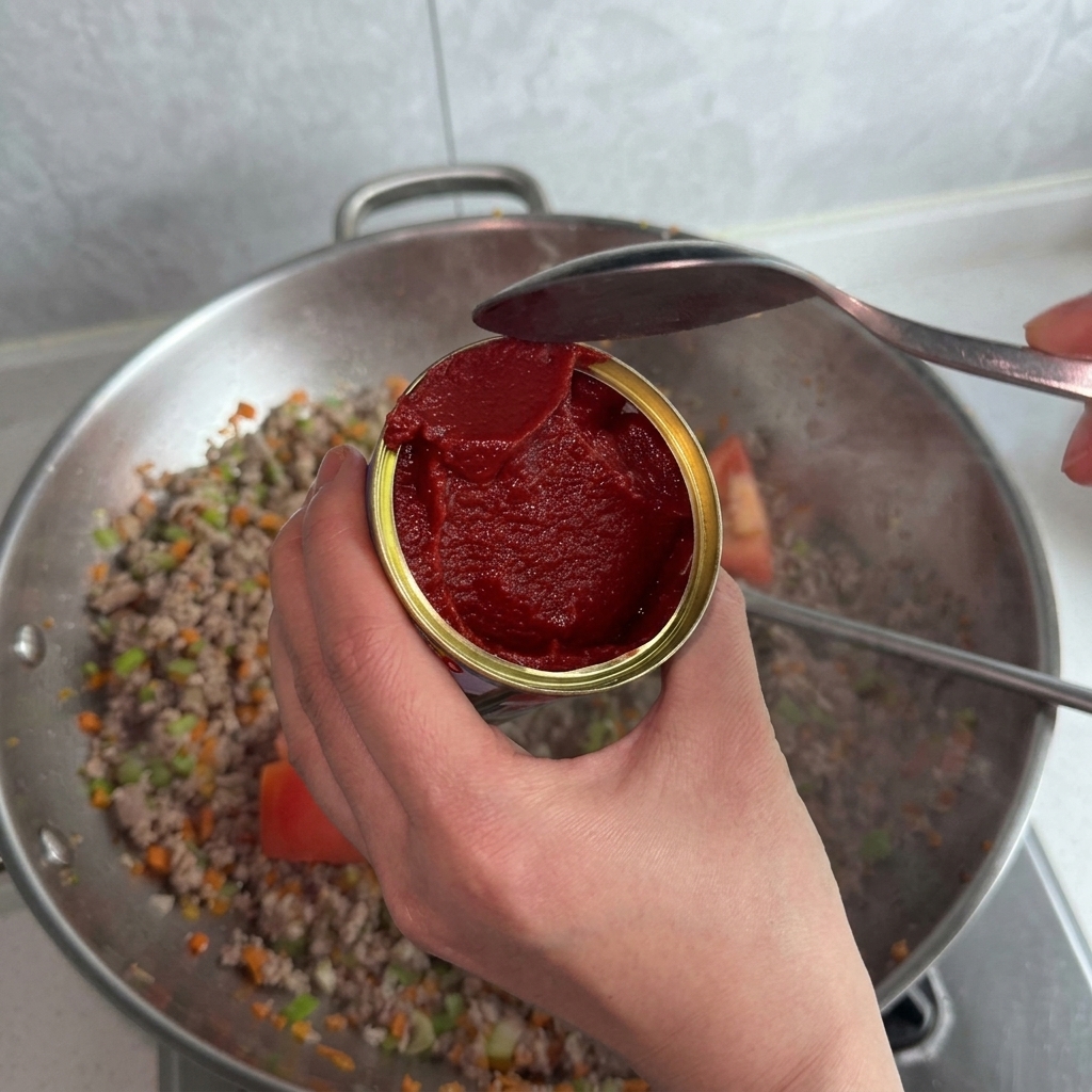 トマトペーストの缶からスプーンで中身をウォック(肉と野菜入り)にすくい入れる様子。