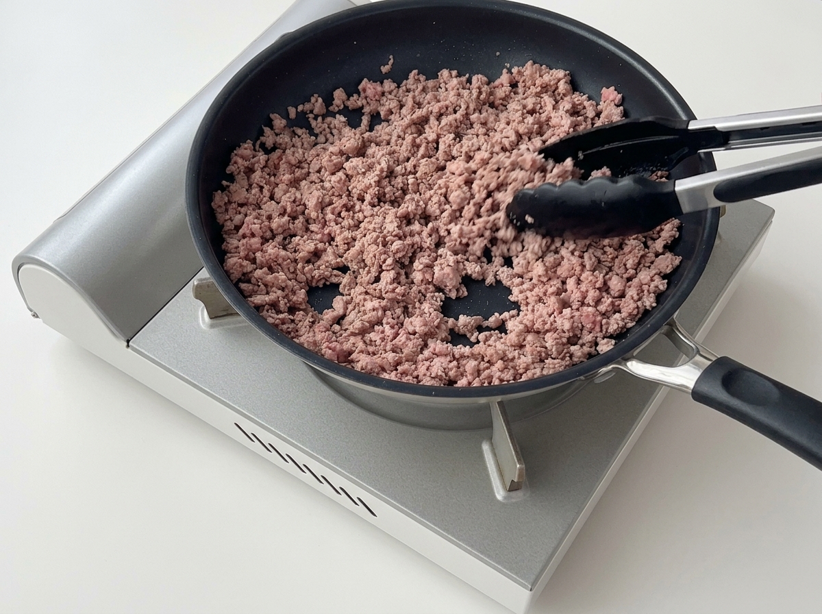 フライパンの中で、トングを使って茶色く色づいたひき肉をほぐしながら炒めている様子。