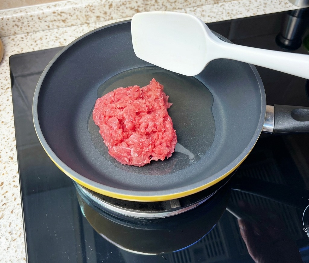 熱したフライパンに油を引き、牛挽き肉の塊をヘラで入れようとしている様子。