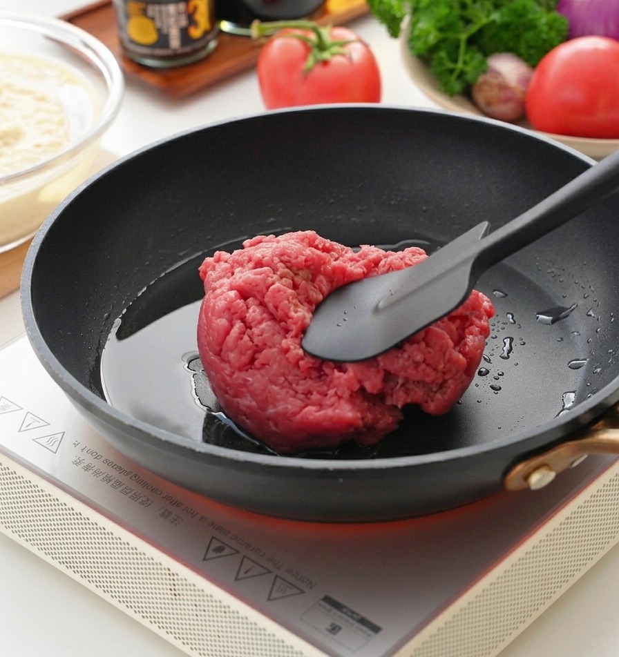 熱した黒いフライパンの上で、ヘラを使って生の牛ひき肉をほぐしている様子。