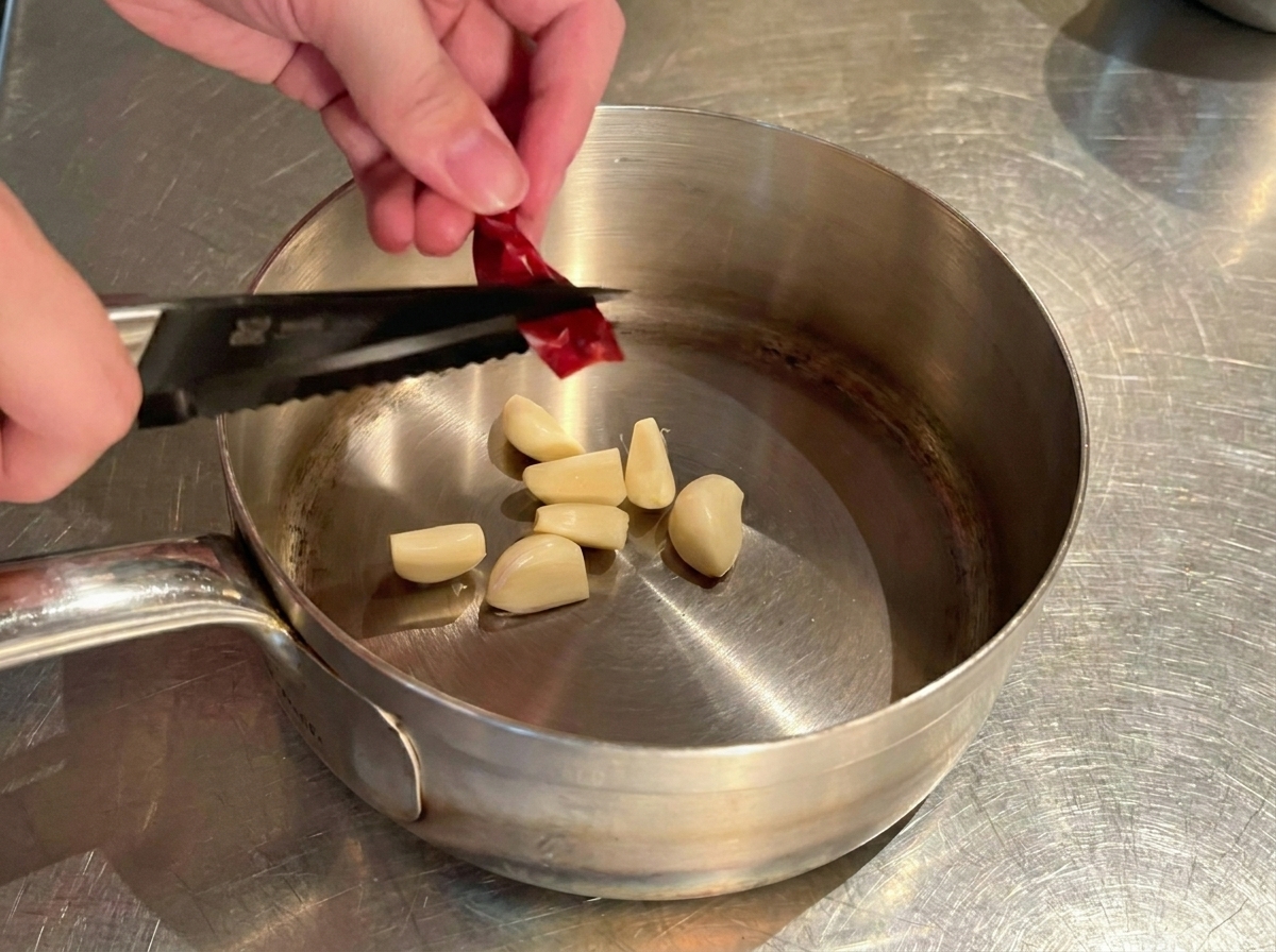 にんにくの入った鍋の上で、キッチンバサミを使って乾燥唐辛子をカットしている手元。