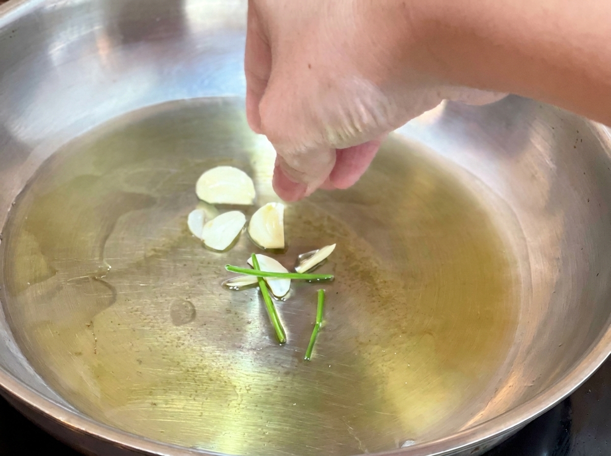 熱いオリーブオイルが入ったフライパンに、スライスしたにんにくとパセリの茎を入れている手元。