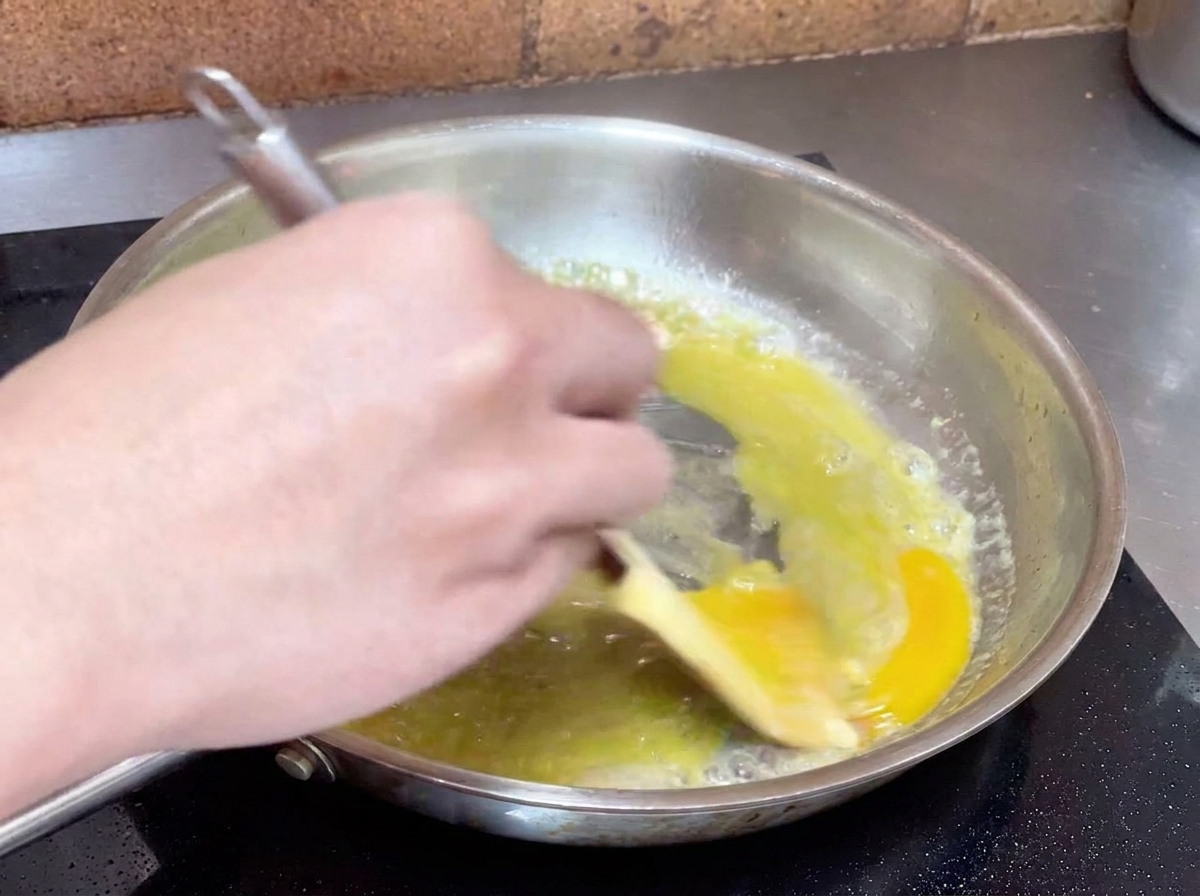 フライパンの中でオイルとゆで汁が混ざり、ヘラで激しくかき混ぜて白っぽいソースを作っている様子。