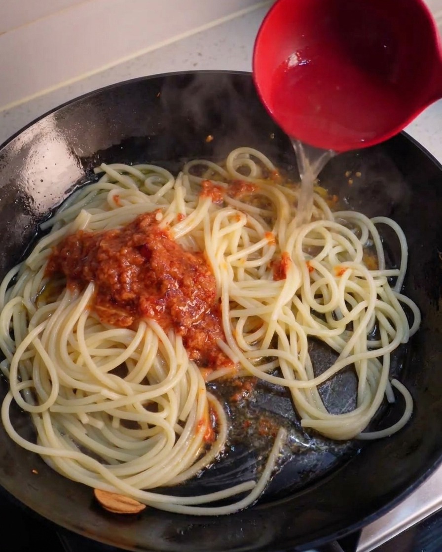 フライパンのスパゲッティに、お玉でパスタのゆで汁とペースト状のソースを加えている様子。