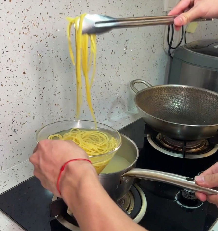トングで茹で上がったパスタを鍋から引き上げ、冷水の入ったガラスボウルに直接移している様子。