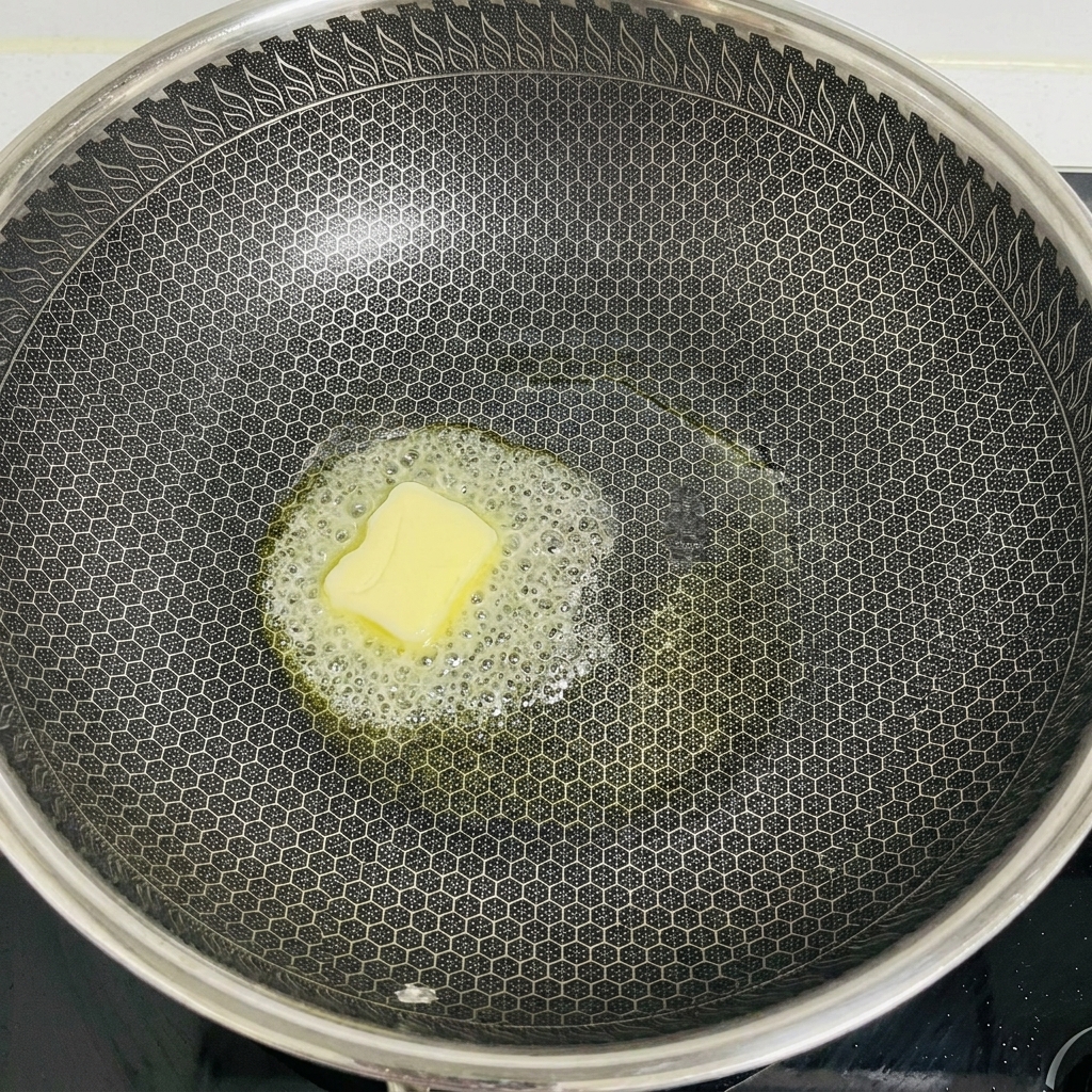ハニカム構造の大きな中華鍋の中央で、四角いバターが泡を立てて溶けている様子。