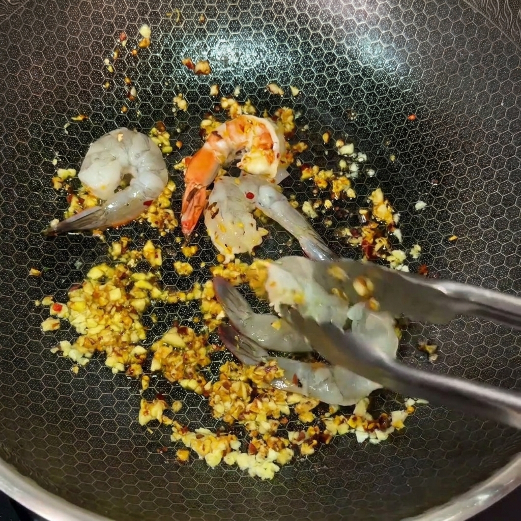 トングを使って、にんにくと唐辛子のバターが熱せられたフライパンに生の海老を並べている様子。