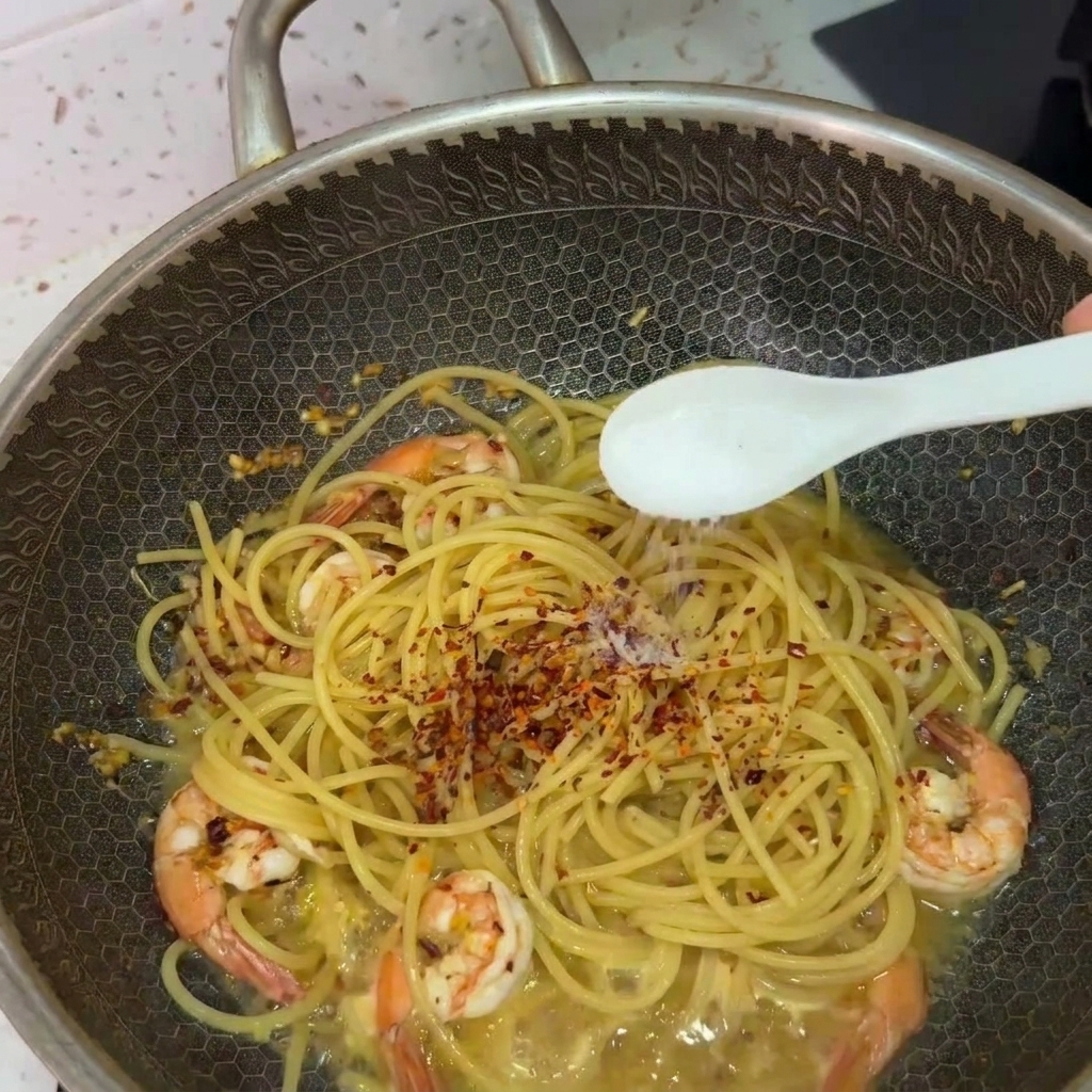 フライパンの中のパスタに白いスプーンで塩を振り、全体を混ぜ合わせている様子。