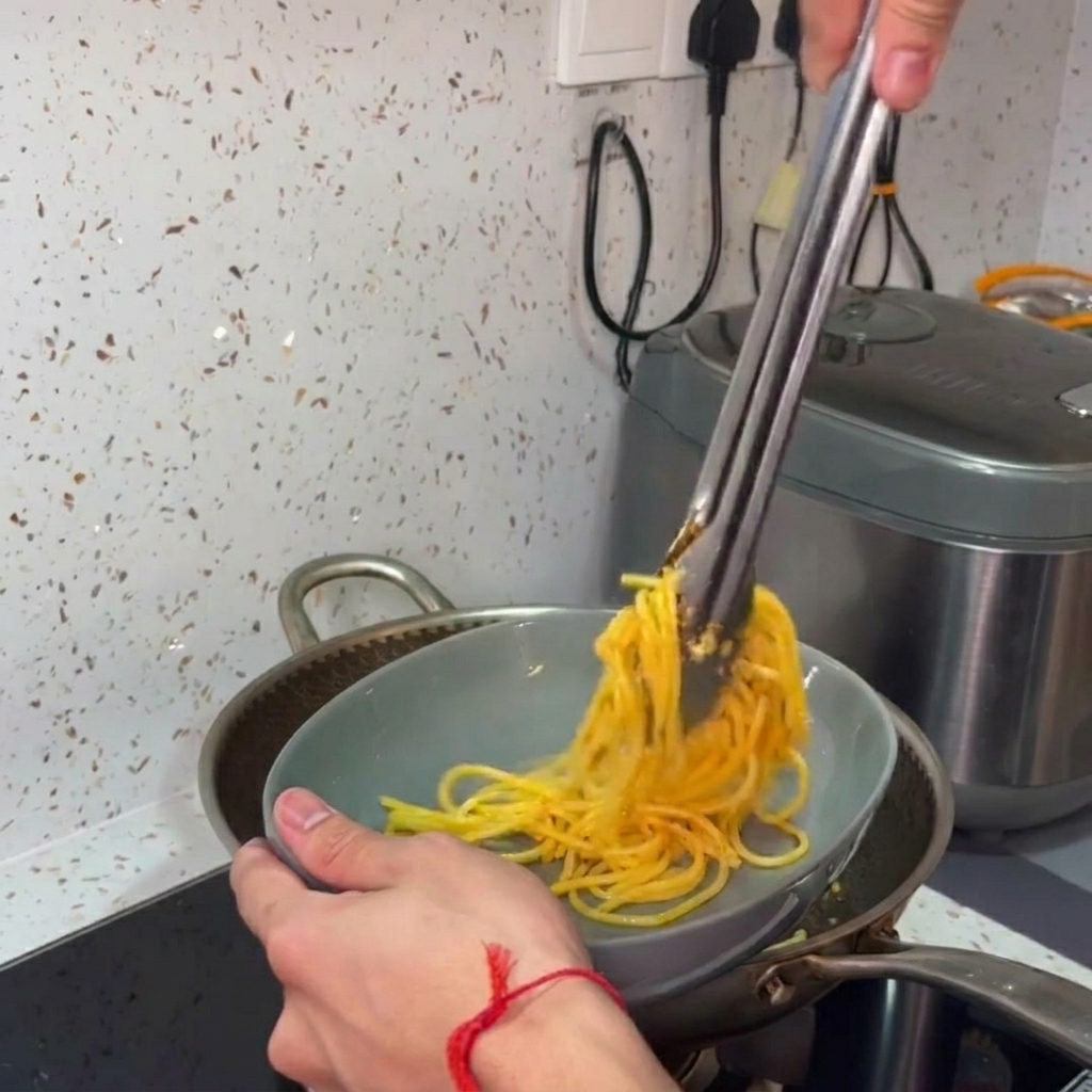 トングを使って、フライパンからパスタをひねりながらグレーの皿に盛り付けている手。